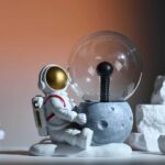 Adorable astronaut figurine sitting next to a vintage-style light bulb on a textured base, perfect for science-themed decor and educational displays.