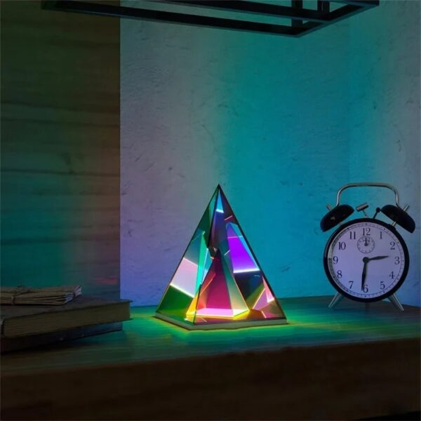 Illuminated geometric pyramid desk lamp with colorful LED lights, placed next to a classic alarm clock and a stack of books on a wooden surface.