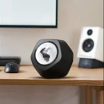 A close-up of a modern, black, geometric-shaped speaker with a unique, illuminated logo on a wooden desk, surrounded by computer accessories and a monitor in a home office setting.