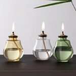 Three glass votive oil lamps with lit wicks, in gold, clear, and green, placed on a black surface against a plain background.