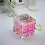 Transparent cube prism with pink and iridescent hues, showcasing light refraction and color effects, placed on a white surface with decorative items in the background.