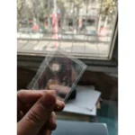 Miniature transparent plastic slide with a tiny circular object inside, held up against a window with a cityscape background.