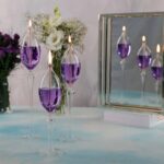 Beautiful purple glass candle holders with lit candles, arranged on a light-colored surface with a floral background and a mirrored display, ideal for science-themed decor and educational settings.