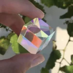 Hand holding a transparent crystal cube with rainbow reflections, showcasing its faceted design against a blurred green leafy background.