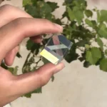 A clear, faceted glass prism held between fingers against a background of green leaves.
