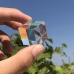 Hand holding a colorful glass cube prism against a clear blue sky, showcasing its reflective and refractive properties.