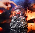 A hand holding a glass crystal ball above a detailed skull sculpture with multiple skulls, set against a fiery, volcanic background.