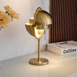 Gold desk lamp with a unique, modern design featuring a spherical shade and elegant curves, placed on a white surface next to a stack of books and flowers.