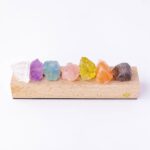 Assorted raw crystals in various colors including clear, purple, blue, pink, yellow, orange, and brown, displayed on a wooden stand against a white background.
