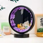 Round mirror with a black frame decorated with pink amethyst crystals, placed on a white surface with decorative items around.