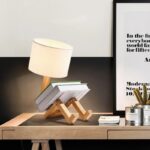 Creative desk lamp made from wooden parts holding books, with a modern design and a white lampshade, placed on a wooden desk next to a framed quote and a small cactus plant.