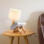 A creative lamp designed as a robot reading a book, placed on a wooden side table next to a cozy armchair. The lamp features a white lampshade and wooden robot legs, adding a playful touch to the room.