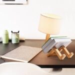 Desk lamp with a wooden stand holding open books, on a modern desk with a beige textured notebook, green water bottle, and black pen holder.