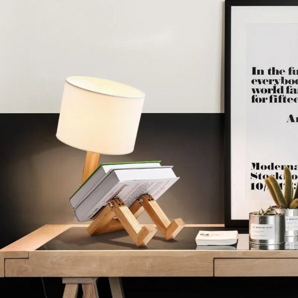 Creative desk lamp made from wooden parts holding books, with a modern design and a white lampshade, placed on a wooden desk next to a framed quote and a small cactus plant.