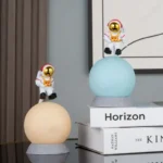 Two astronaut cat figurines with gold helmets sitting on illuminated spheres, displayed on books in a modern interior setting.
