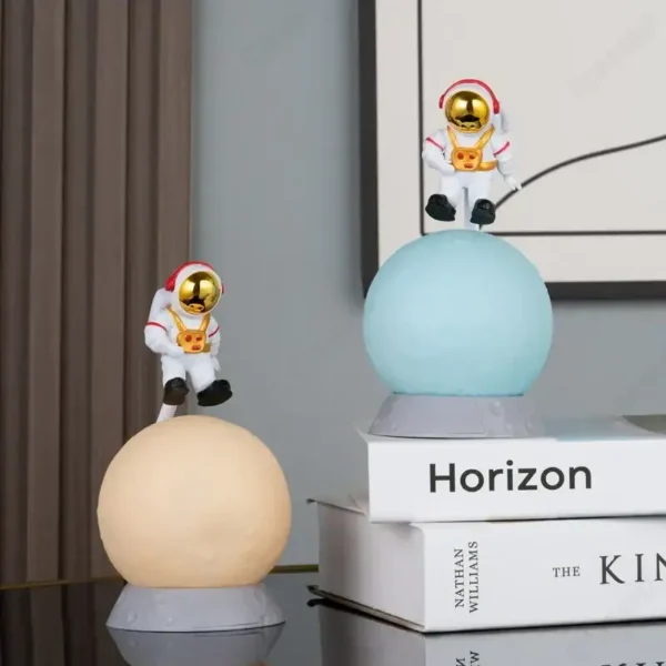 Two astronaut cat figurines with gold helmets sitting on illuminated spheres, displayed on books in a modern interior setting.