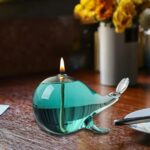 Glass ocean wave-shaped candle lamp with a lit flame, placed on a wooden surface with a blurred background of flowers in a white vase.