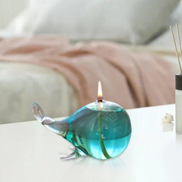 Elegant whale-shaped glass oil lamp with a small flame on top, placed on a white surface in a cozy indoor setting.