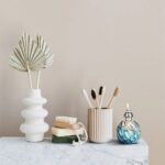Stylish bathroom setup featuring toothbrushes in a striped holder, a white textured vase with dried leaves, a soap dish, and a decorative glass oil diffuser on a marble surface.