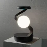 A modern decorative light sculpture featuring a black spiral stand with a glowing white sphere at its center, placed on a white pedestal against a textured gray wall.