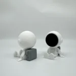 Two minimalist astronaut figurines with spherical heads, one with a black faceplate and the other with a plain white head, sitting on a white surface.