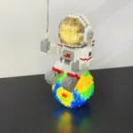 LEGO astronaut figurine sitting on a vibrant, rainbow-colored base with a rainbow pattern, featuring a white space suit with gray and red details, and a gold visor helmet.