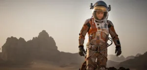 An astronaut in a detailed space suit walking across a rocky, desert-like terrain with mountains in the background under a hazy sky.