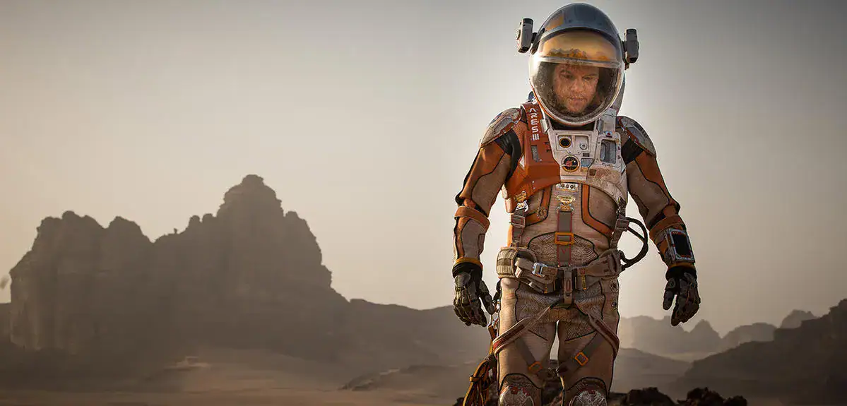 An astronaut in a detailed space suit walking across a rocky, desert-like terrain with mountains in the background under a hazy sky.