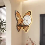 A decorative butterfly-shaped wall clock featuring a white clock face with black and red hands, mounted on a beige wall. The clock has a warm LED backlight highlighting its butterfly design.
