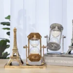 A decorative Eiffel Tower miniature with a vintage-style hourglass in the center, placed on a wooden surface alongside a small replica of the Eiffel Tower.
