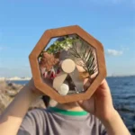 Hand holding a hexagonal glass terrarium with seashells, green leaves, and decorative stones, set against a seaside background with blue sky and water.