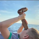 Child playing with a wooden telescope by the sea, exploring the horizon during daytime.