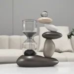 Modern decorative sculpture featuring stacked smooth stones and a glass sand timer, placed on a white surface in a contemporary living room setting.