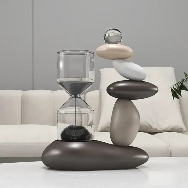 Modern decorative sculpture featuring stacked smooth stones and a glass sand timer, placed on a white surface in a contemporary living room setting.