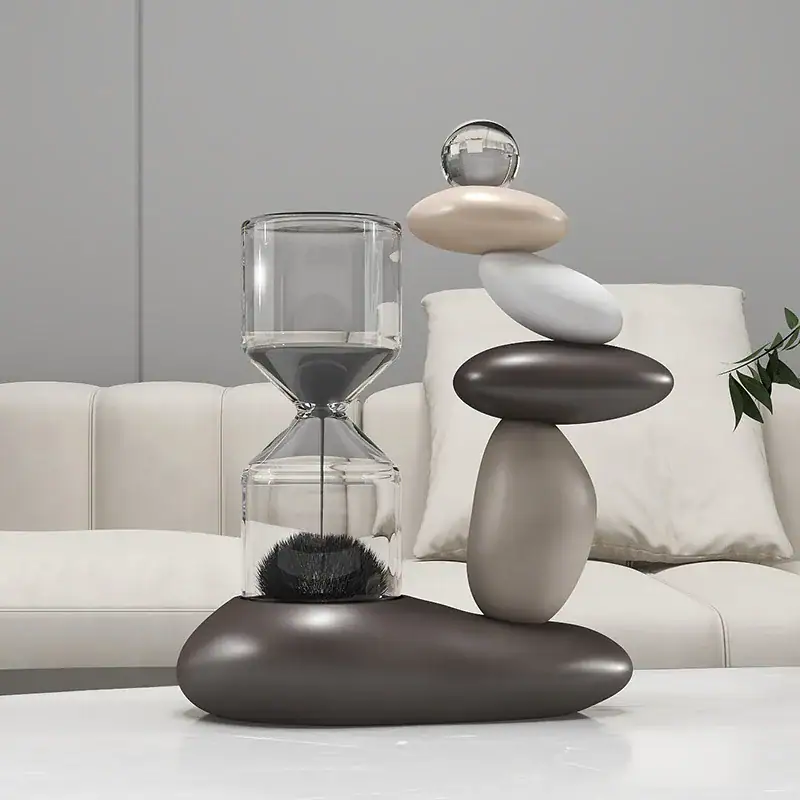 Modern decorative sculpture featuring stacked smooth stones and a glass sand timer, placed on a white surface in a contemporary living room setting.