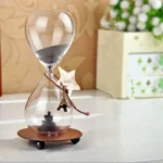 Stylish hourglass with black sand, mounted on a wooden base, featuring a small Eiffel Tower charm hanging from the top. Perfect for home decor or educational purposes.