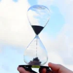 A close-up of a classic glass hourglass timer with black sand, demonstrating the passage of time, held outdoors with a clear sky in the background.