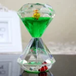 Transparent hourglass-shaped liquid timer with green liquid, featuring a yellow ladybug and a red toy at the bottom, used for educational and decorative purposes.