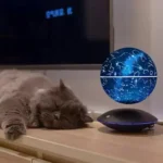 A star map globe displaying constellations and celestial patterns, placed on a sleek black base, with a relaxed cat sleeping beside it on a wooden surface.