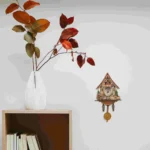 Autumn leaves in a white ceramic vase placed on a wooden shelf, with a decorative cuckoo clock on the wall in the background.