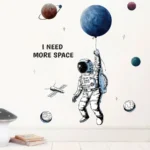 Whimsical space-themed wall art featuring an astronaut holding a planet balloon surrounded by planets and stars, with the phrase "I NEED MORE SPACE" in bold letters.