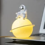 Astronaut lamp sitting on a yellow planet-shaped base with a ring, illuminated softly, creating a space-themed decorative piece. Perfect for science enthusiasts and educational settings.