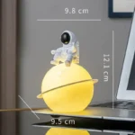Cute astronaut lamp sitting on a yellow planet-shaped night light with ring, measuring 9.8 cm in height and 9.5 cm in diameter, perfect for science-themed decor.