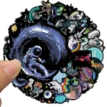 Hand holding a sticker of an astronaut floating in space with a swirling galaxy background, part of a collection of science-themed stickers.