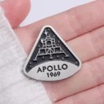 Close-up of a metallic Apollo 11 badge featuring the lunar module and the inscription "APOLLO 1969" held in a person's hand.