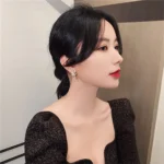 Woman with black hair styled in a low bun, wearing a black, sparkly dress with puffed sleeves, and red lipstick, posing against a neutral background.