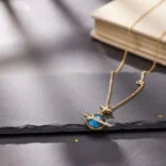Elegant gold chain necklace featuring a star and planet pendant with blue gemstone accents, resting on a dark surface near a closed notebook.