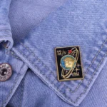 Enamel pin featuring a space-themed design with a rocket and planets, attached to denim fabric.