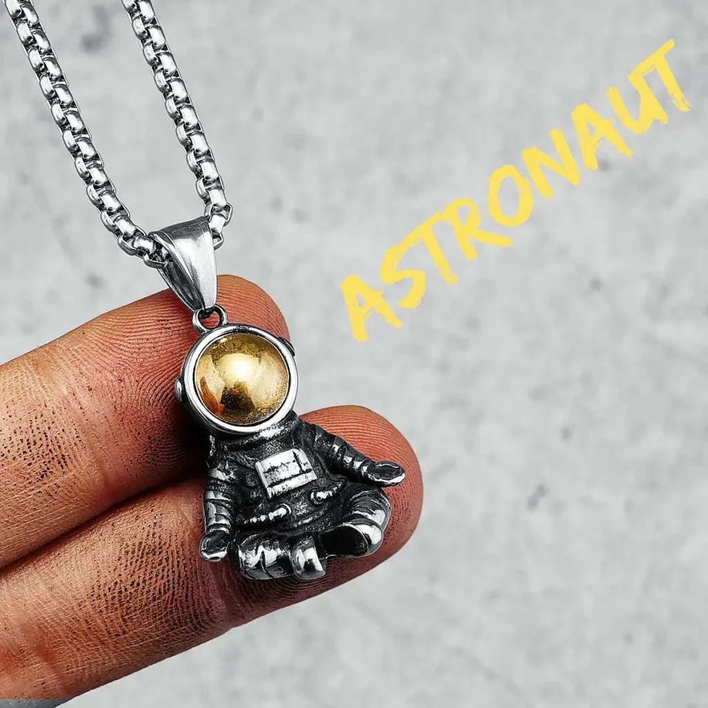 Close-up of a silver astronaut pendant necklace with a gold sphere helmet, held between fingers against a neutral background.
