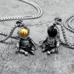 Two astronaut-themed pendant necklaces featuring detailed space suits and spherical helmet designs, one with a gold-colored helmet and the other with a black helmet, on a textured gray background.